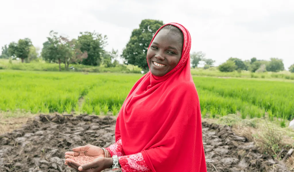 Nigerian woman is smiling in a red hijab, with seeds held in her open palms against lush green fields in the background.
