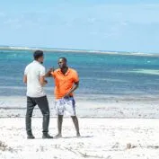 Two men stand on a white sand beach and talk. The ocean is behind them.