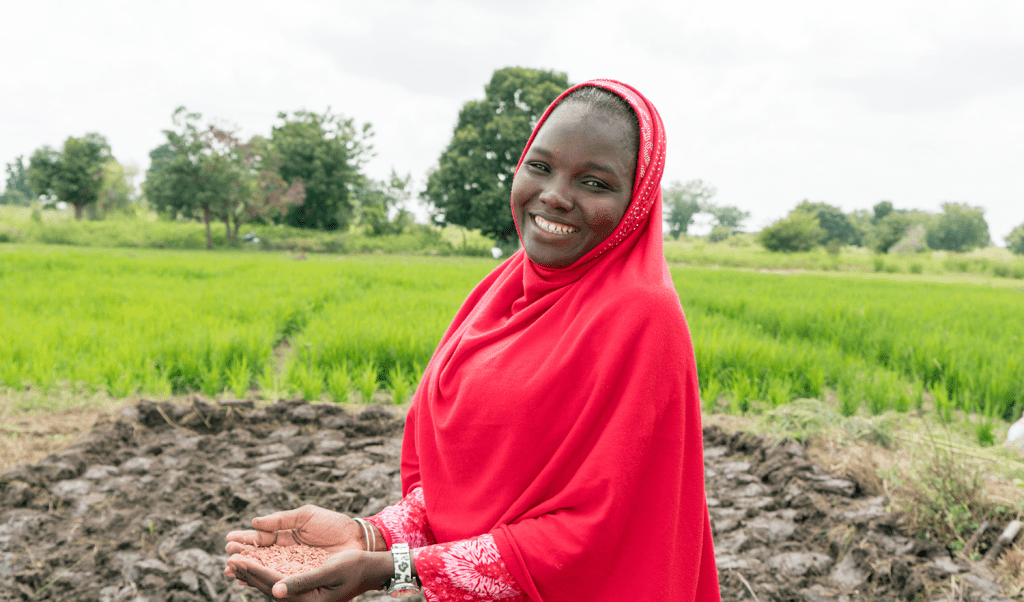 Nigerian woman is smiling in a red hijab, with seeds held in her open palms against lush green fields in the background.