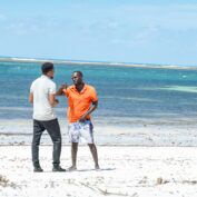 Two men stand on a white sand beach and talk. The ocean is behind them.