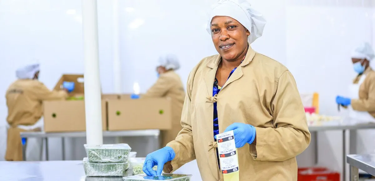 A worker in a food processing facility smiles while handling packaged food items.