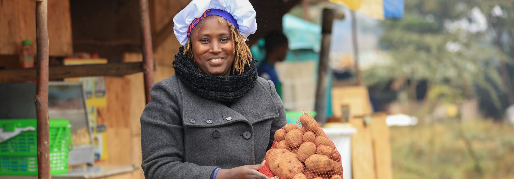 A person smiles while holding a large bag of potatoes at an outdoor market stall.
