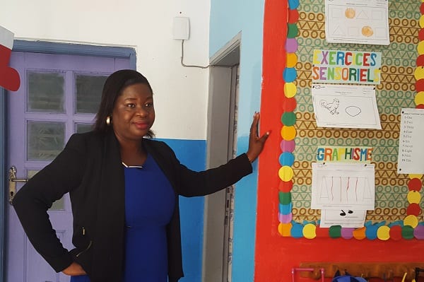 A woman in a blazer and blouse standing up looking forward in a classroom.