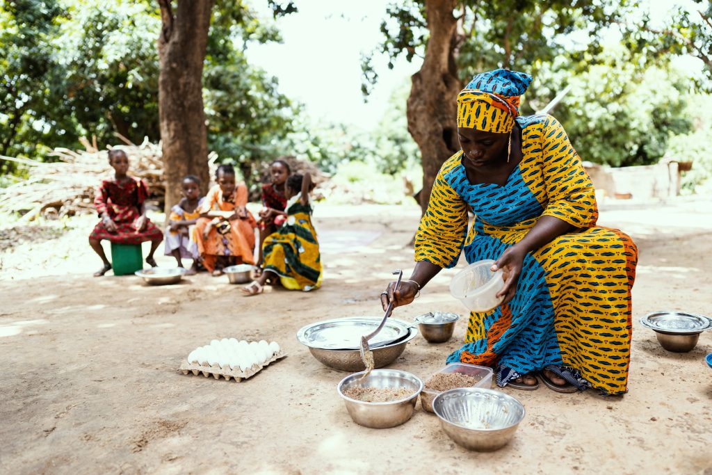 In this image, a colourfully dressed black African mother is preparing a meal for her family, with her daughters waiting in the background