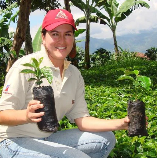 A woman wearing a red cap kneels in a lush green field, holding two young plant seedlings wrapped in black plastic bags. She smiles at the camera with a mountainous landscape in the background.