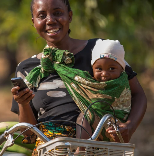 A smiling woman rides a bike, holding a phone, with a baby in a green cloth sling. The baby wears a white hat that says "baby."