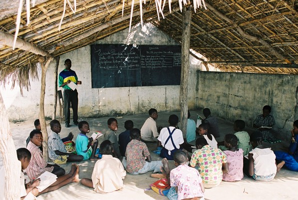 A group of children sitting on the floor in a classroom with a thatched roof, listening to a teacher standing near a blackboard.