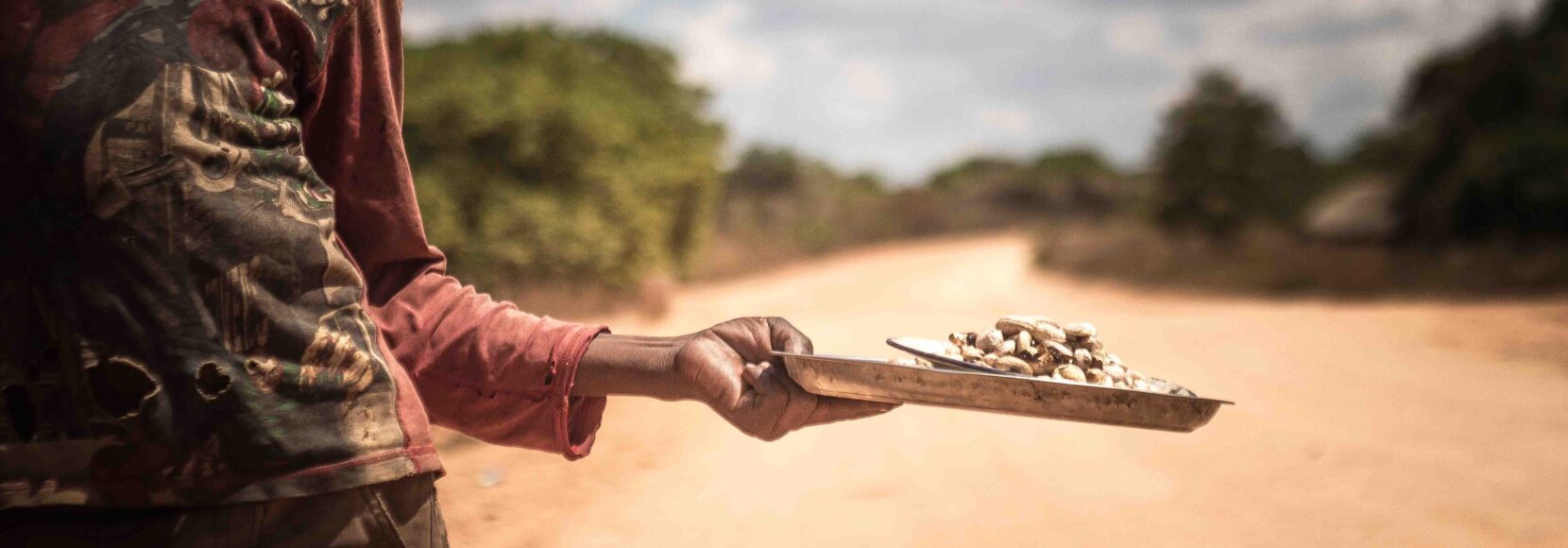 A person holding a tray of cashew extended in one hand, standing on a dirt road under a cloudy sky.
