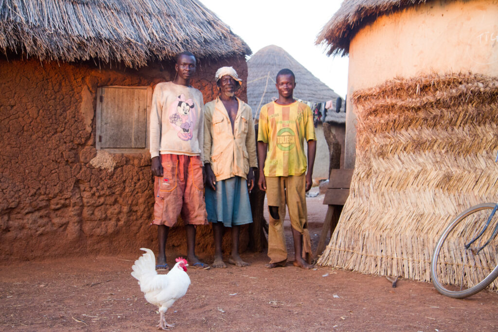 Three men standing outside traditional round mud huts, with a chicken in the foreground.