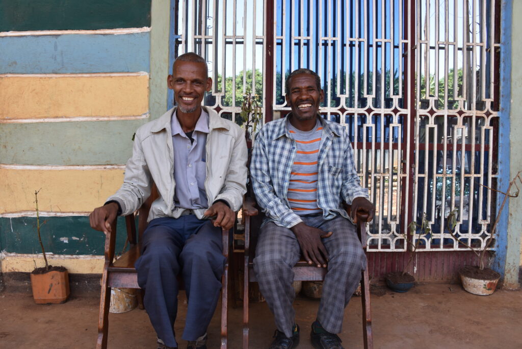 Two men sitting in chairs, smiling outside a building with colorful painted walls and a metal gate.