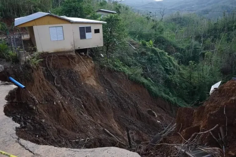 A small house precariously perched at the edge of a large landslide, with much of the surrounding land eroded away.