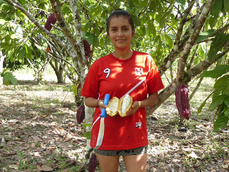 A woman in a soccer jersey holds a cacao pod on her farm in Haunuco, Peru