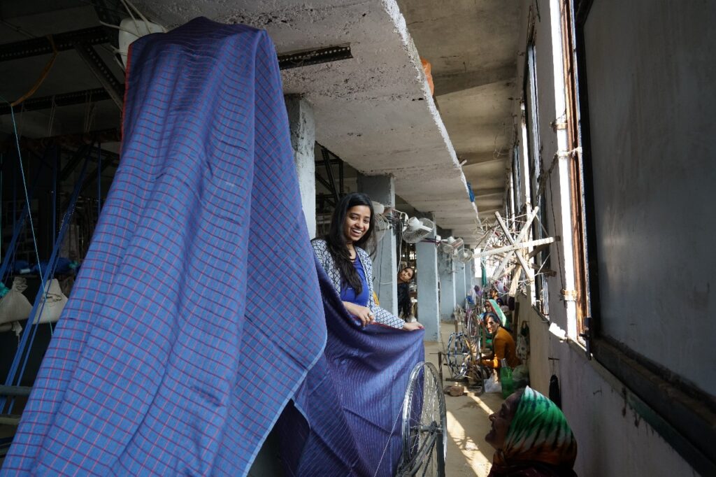 A woman smiles while holding blue fabric and standing in a shop in India.