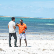 Two men stand on a white sand beach and talk. The ocean is behind them.