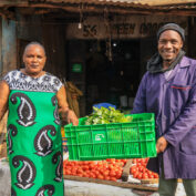 Two individuals stand outside a market stall, holding a crate of fresh greens and surrounded by piles of tomatoes for sale.