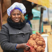 A person smiles while holding a large bag of potatoes at an outdoor market stall.