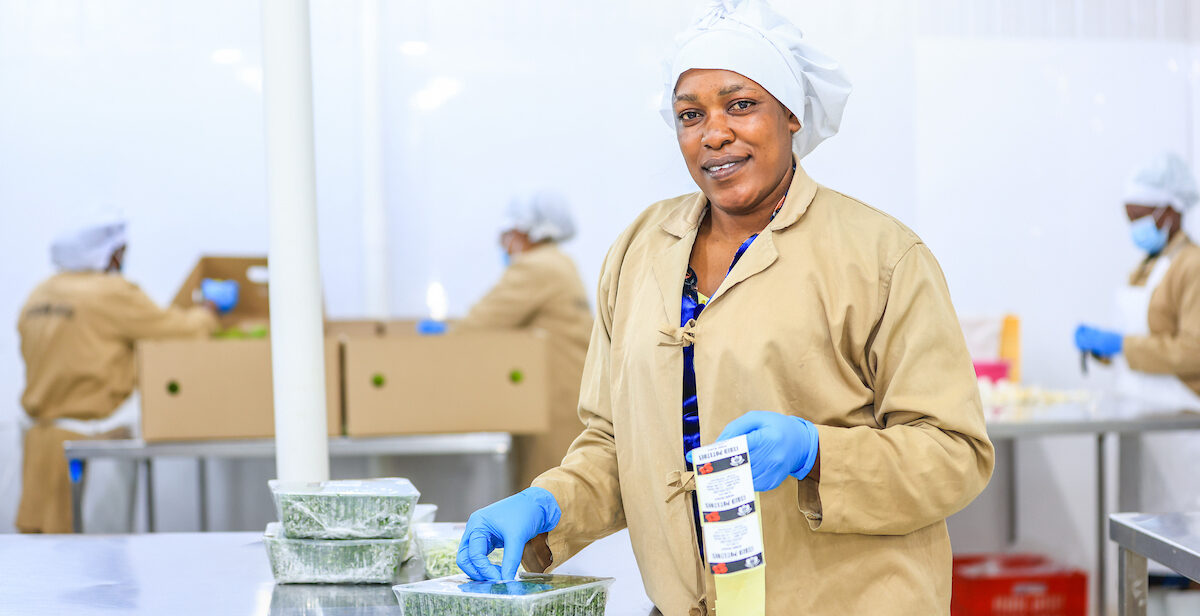 A worker in a food processing facility smiles while handling packaged food items.