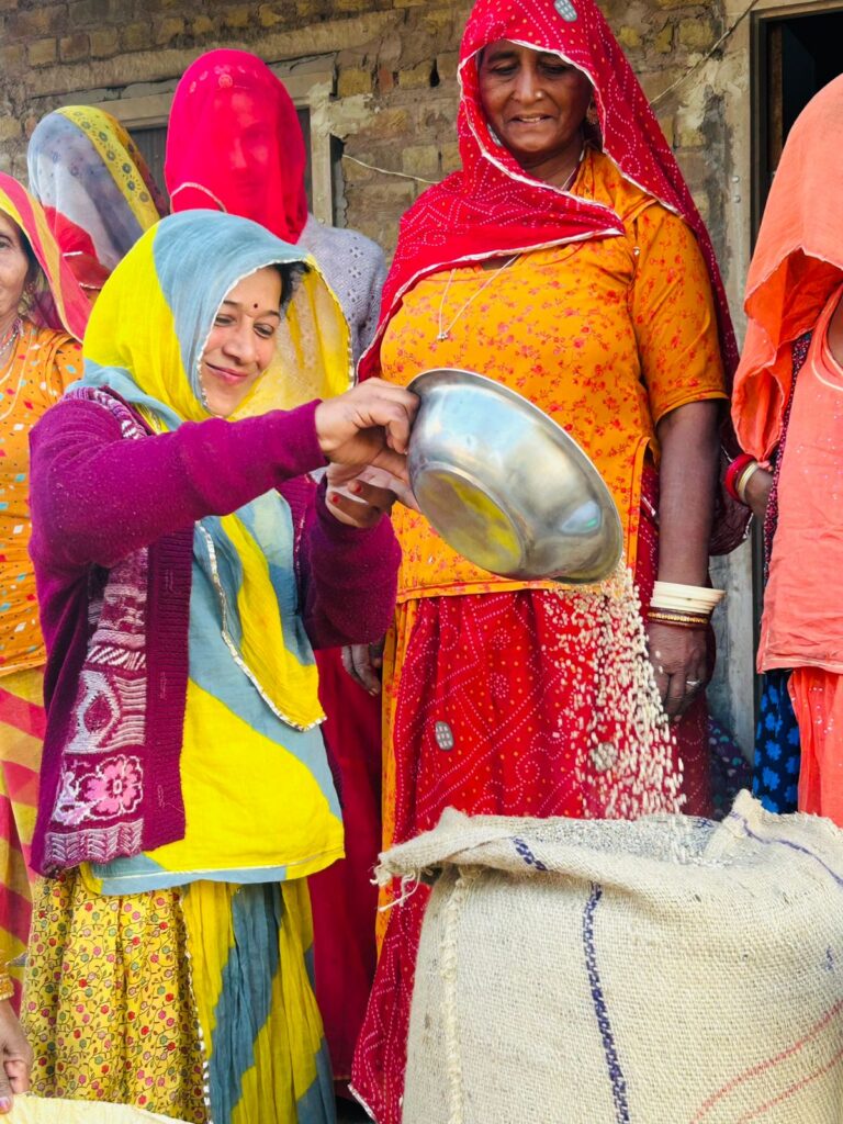 A smiling woman farmer four guar seeds from a shiny aluminium bowl into a gunny sack as other women in colorful clothes look on. She is part of a female farmer group that TechnoServe helped support to gain negotiate better prices as part of the Sustainable Guar Initiative in the Indian state of Rajasthan.